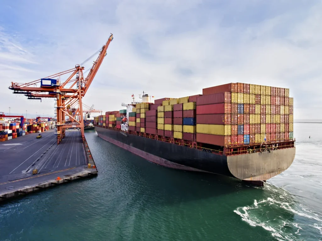Container ship being loaded at a port terminal with large cranes, surrounded by stacked shipping containers.