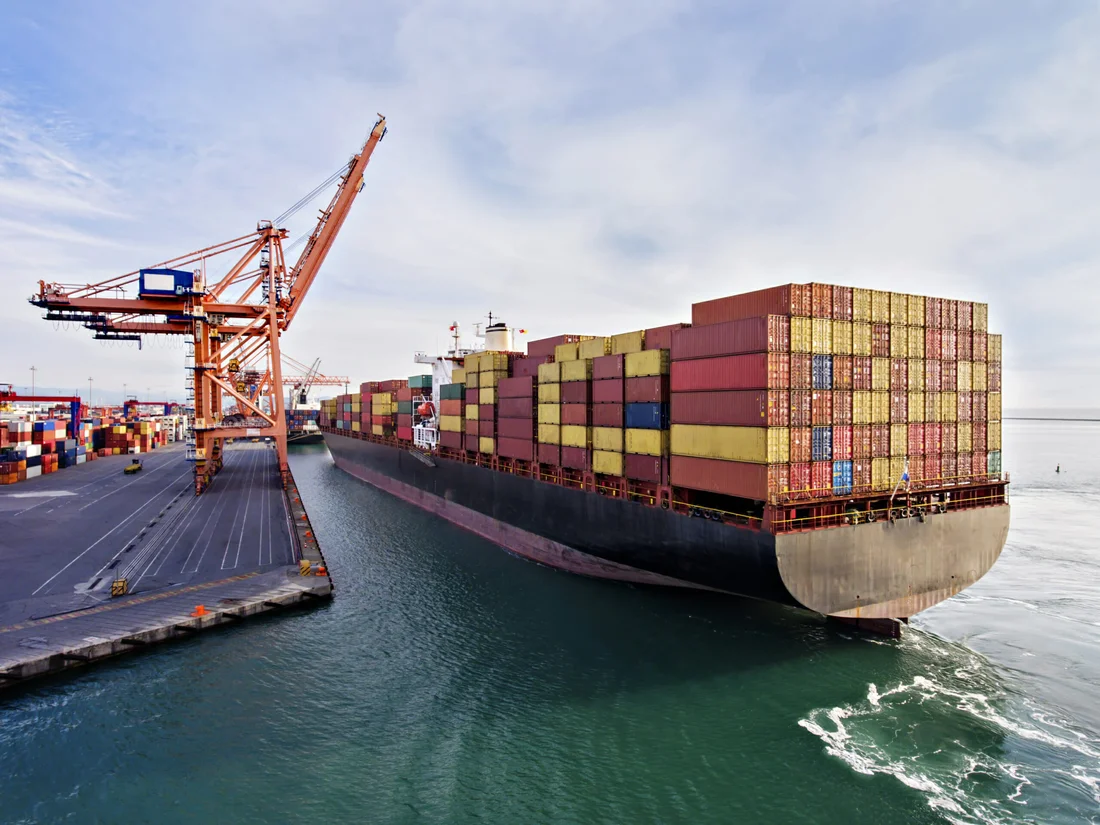 Container ship being loaded at a port terminal with large cranes, surrounded by stacked shipping containers.