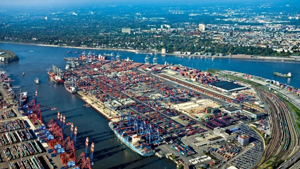 The image shows an expansive aerial view of a busy container port with numerous cranes, cargo containers, and shipping vessels. A nearby urban area and rail connections highlight the integration of maritime and inland logistics.