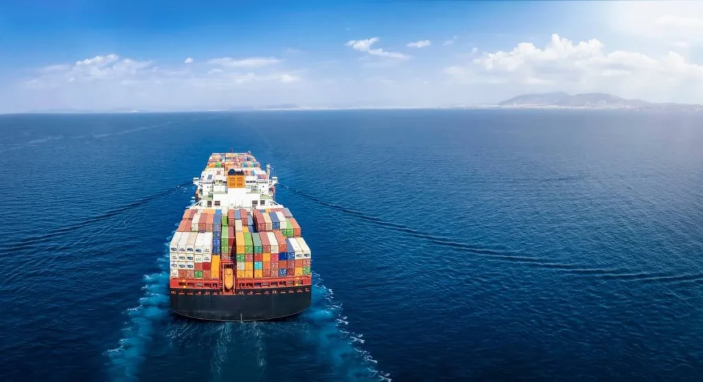 The image shows a large container ship navigating through calm blue seas under a clear sky. The ship is fully loaded with colorful cargo containers, leaving a visible wake in the water.