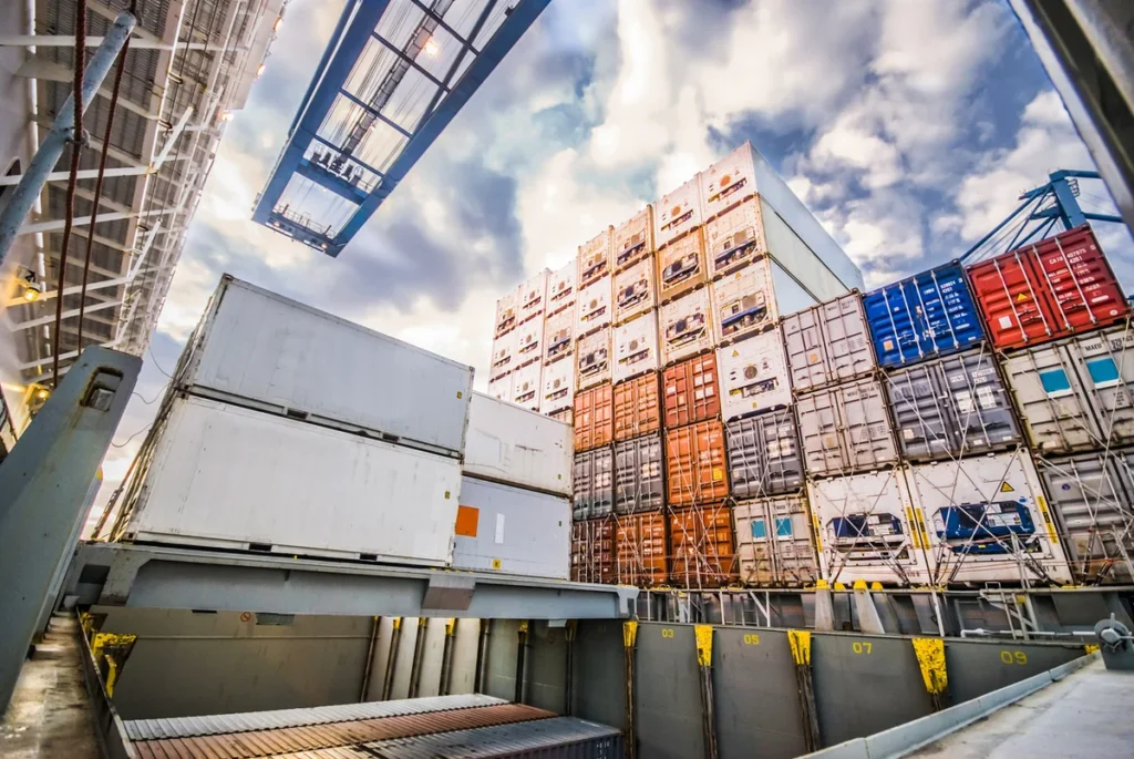 The image shows stacked cargo containers, including refrigerated containers (reefers), on a ship deck, with a crane above under a partly cloudy sky. The containers are securely fastened, ensuring safe transportation.
