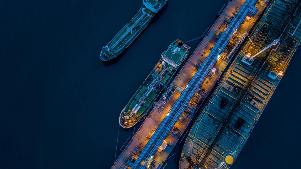 Aerial view of two liquid bulk carrier ships docked at a port, with pipelines and loading equipment visible, illuminated in the evening light.